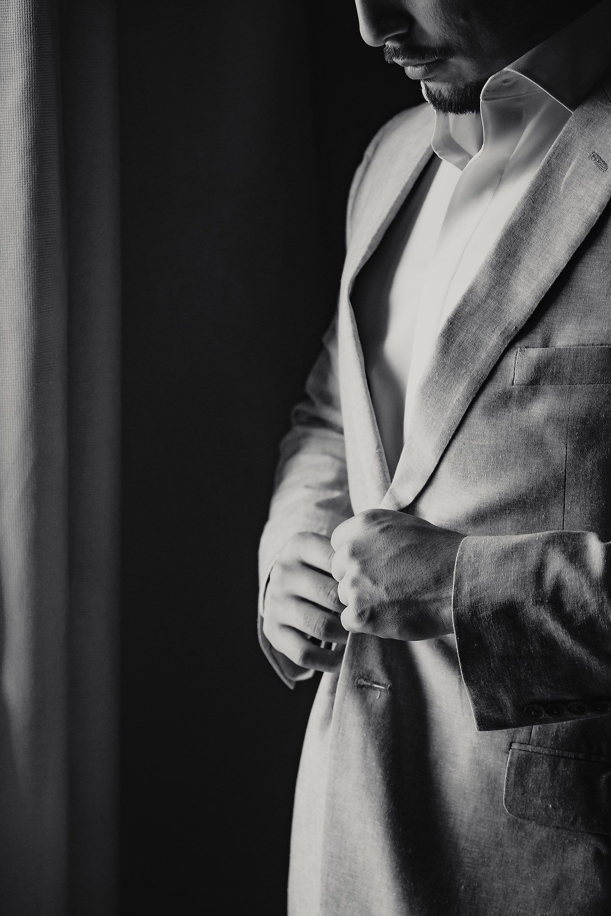 Black and white close-up of groom buttoning grey suit jacket before wedding ceremony.