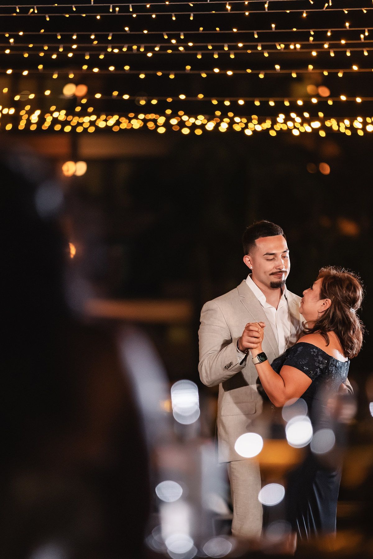 Groom dances with his mother under warm string lights at an elegant outdoor wedding reception at night.