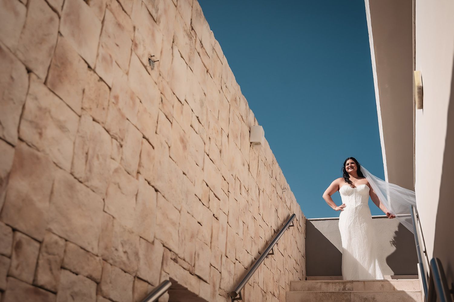 Bride in white gown poses on stone staircase against sandy brick wall and blue sky.