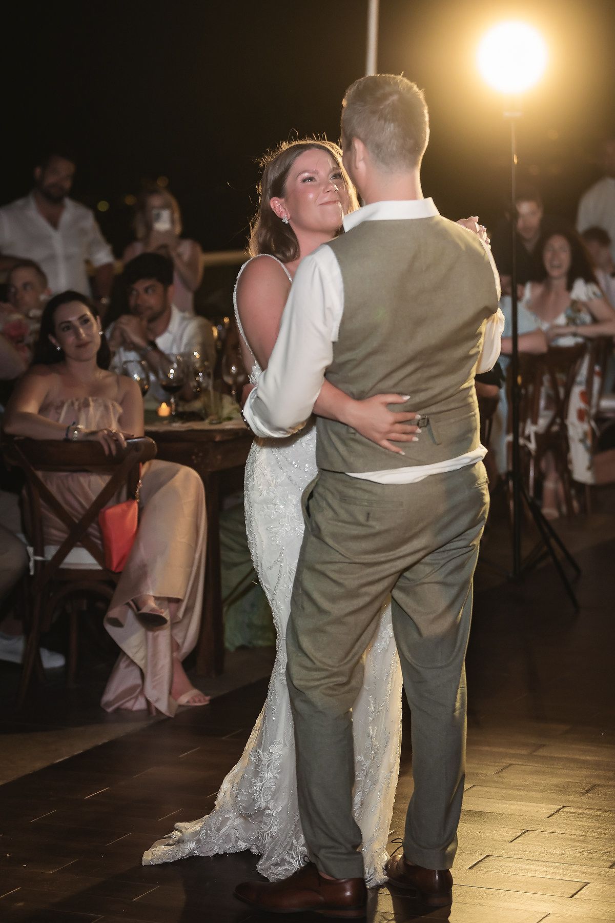 Bride and groom share their first dance at an outdoor wedding reception, surrounded by guests under warm evening lights.