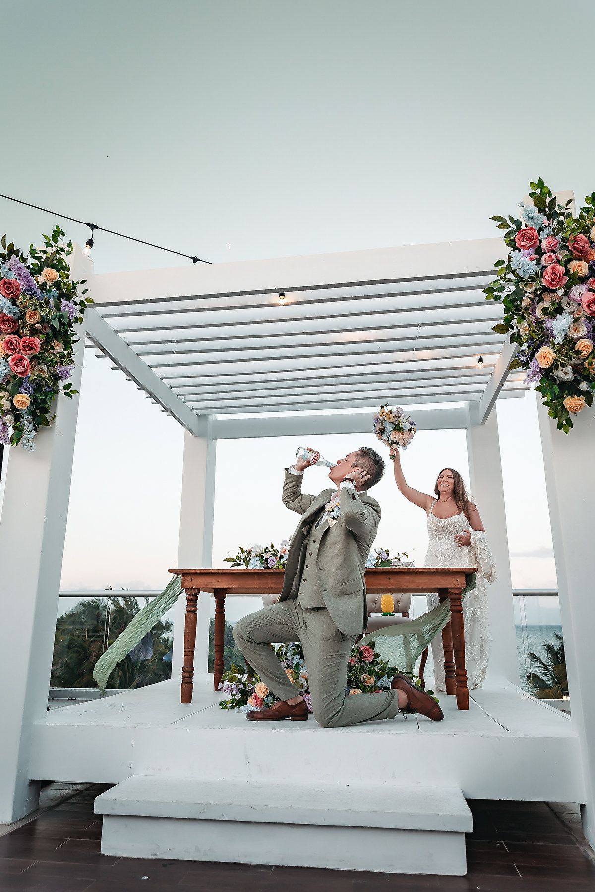 Groom kneeling and drinking at outdoor wedding ceremony under floral pergola while bride holds bouquet.