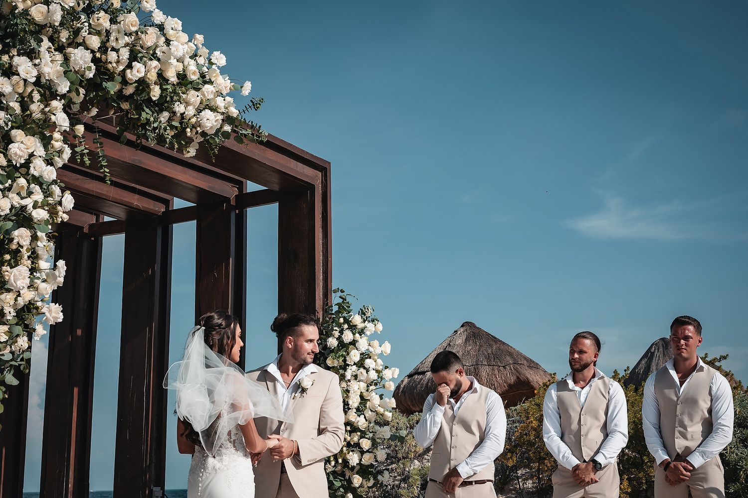 Outdoor beach wedding ceremony with bride and groom at floral arch, groomsmen in tan suits under blue sky.