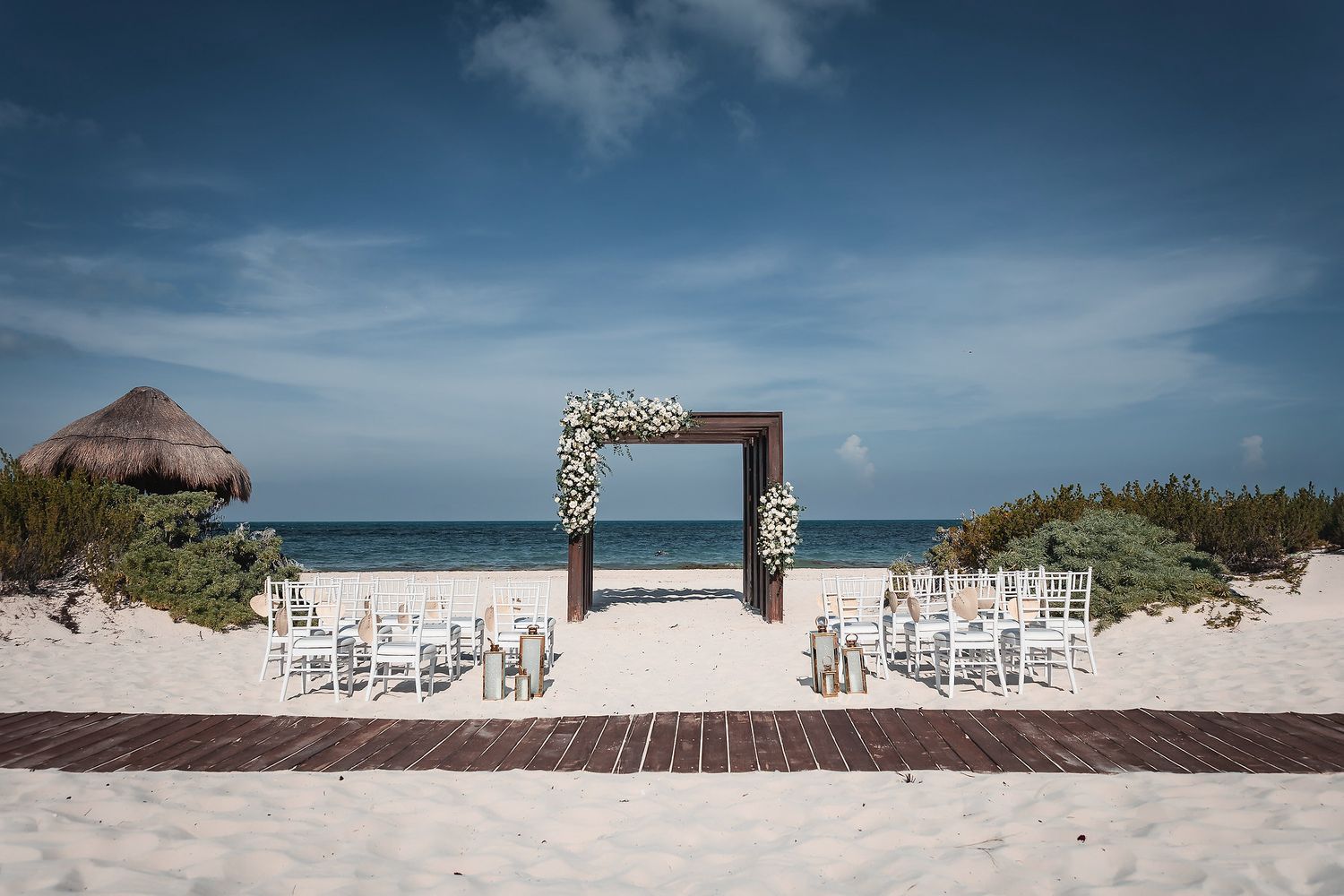 Beach wedding ceremony setup with floral arch, white chairs, and ocean backdrop under blue sky.