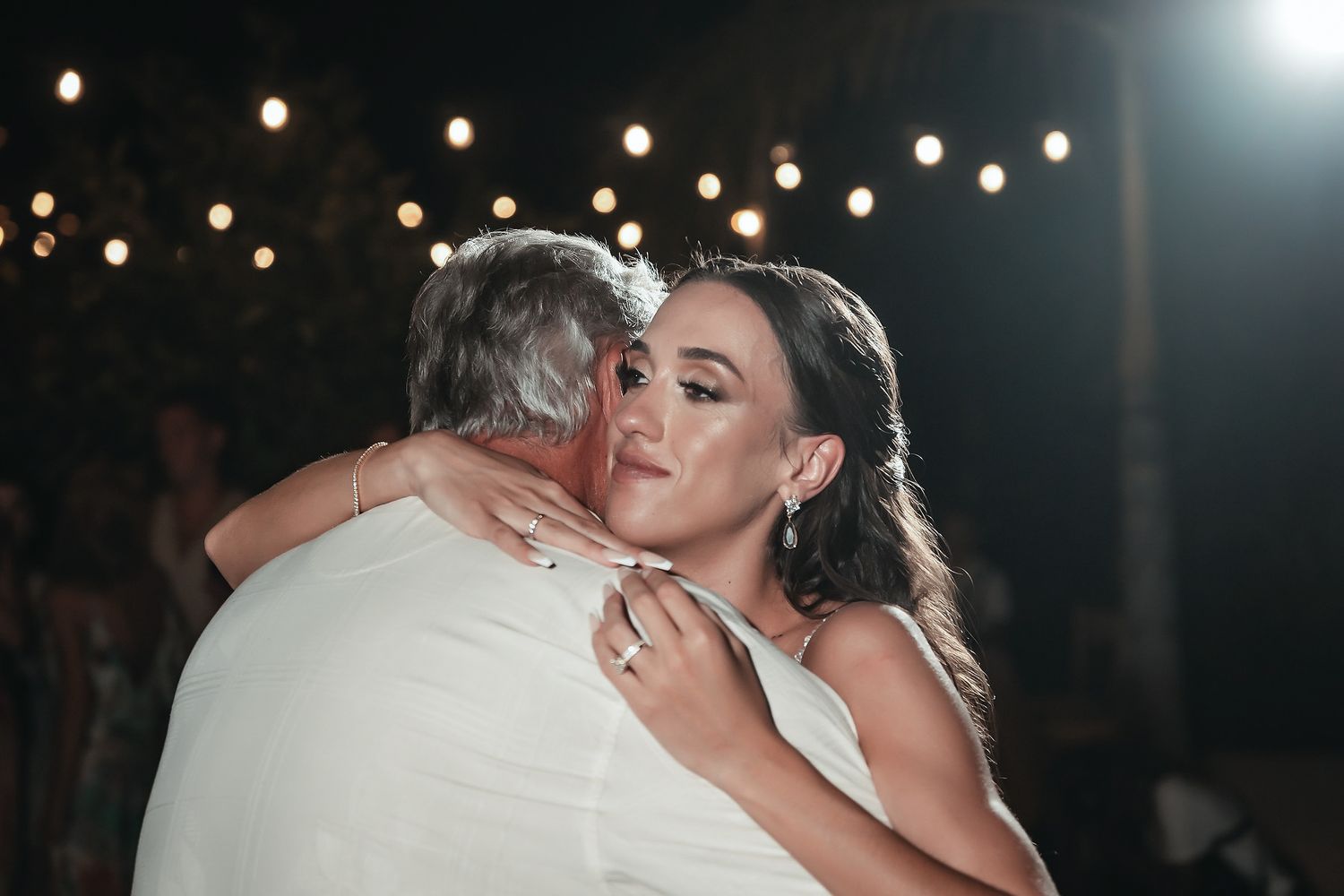 Bride and father share emotional first dance embrace under string lights at outdoor wedding reception.
