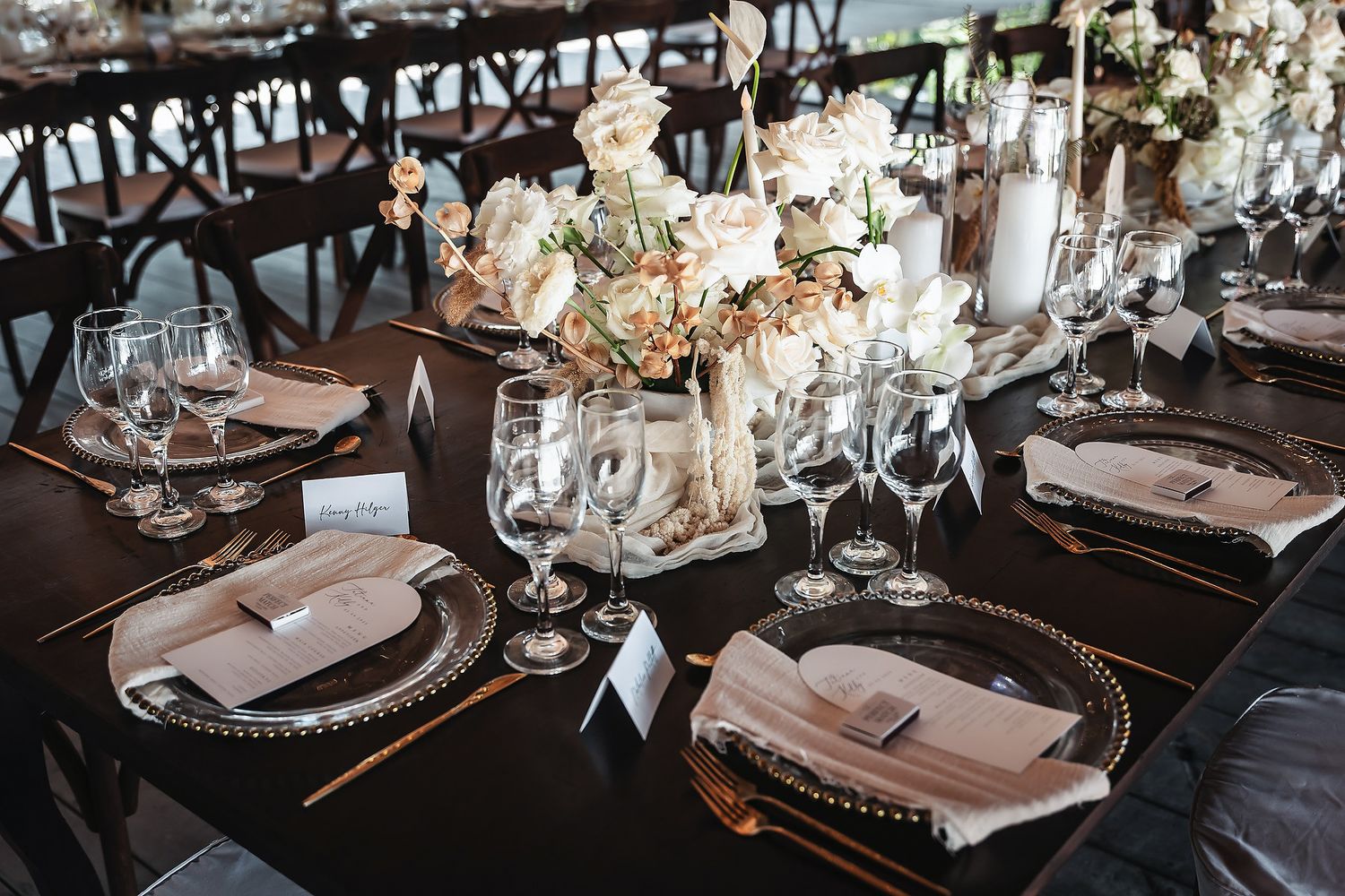 Elegant wedding reception table with gold charger plates, crystal glasses, and blush floral centerpieces on dark wood tables.