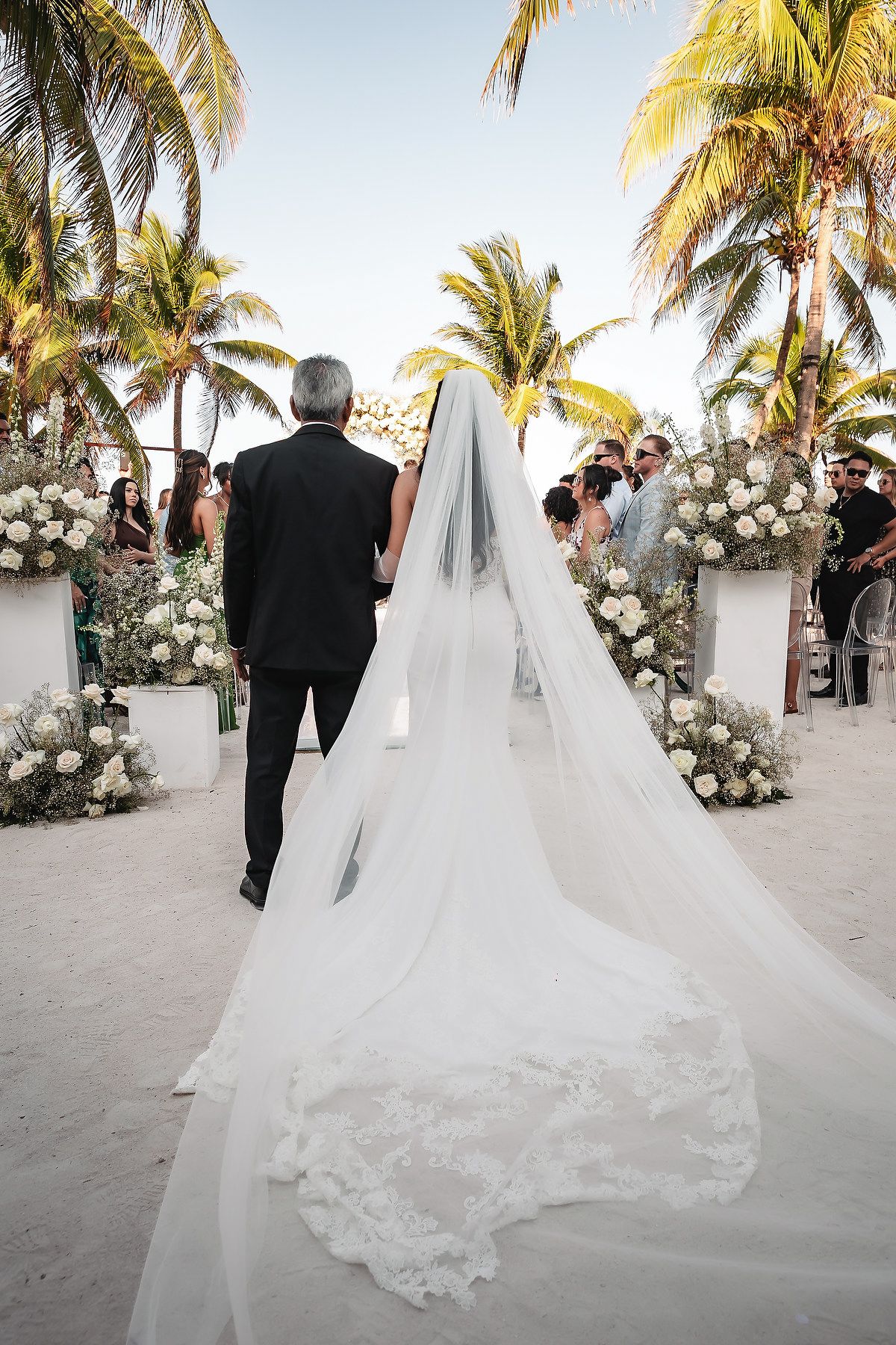 Bride with long lace-trimmed cathedral veil walks toward floral arch on tropical beach ceremony.