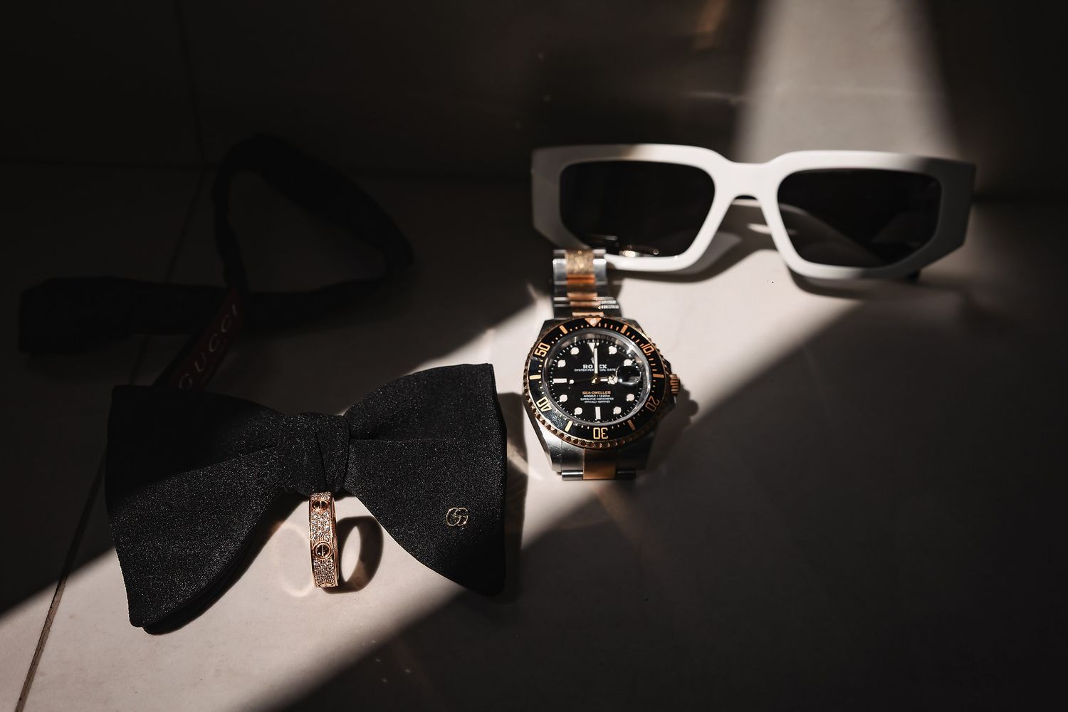 Luxury men's accessories flat lay featuring a Rolex watch, white sunglasses, bow tie, and gold ring in dramatic lighting.
