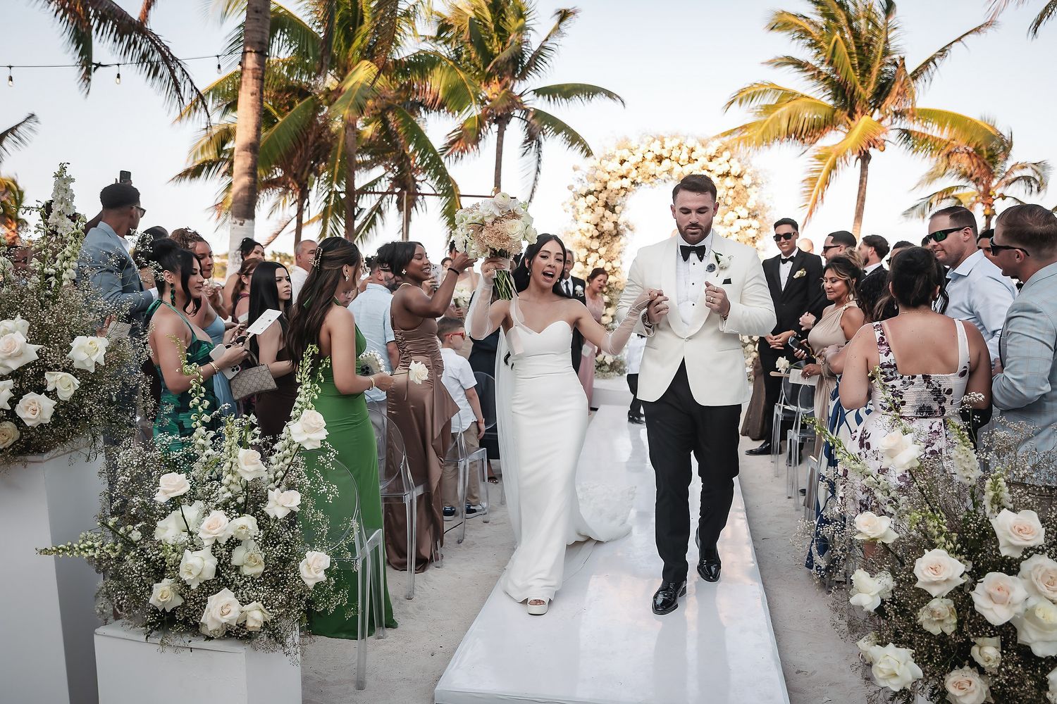 Bride and groom exit wedding ceremony on white aisle with white floral arrangements and tropical palm trees.
