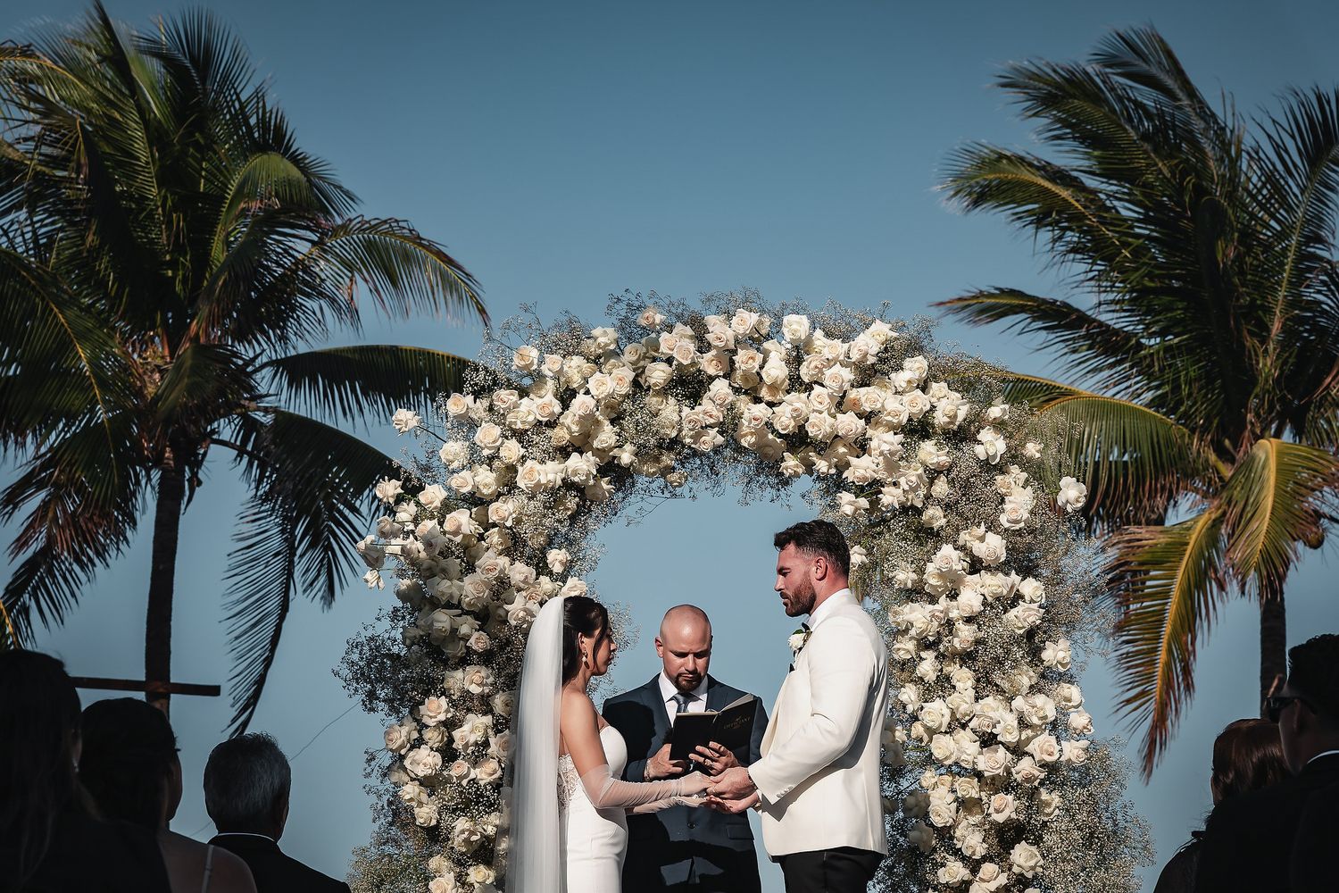 Couple exchanges vows under a stunning white floral arch with palm trees and blue sky at a tropical beach wedding.
