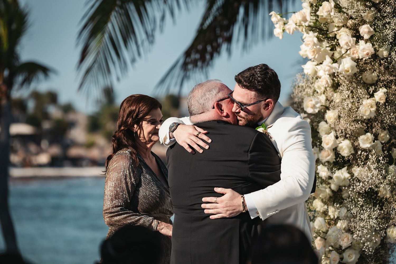 Groom embracing guests at outdoor beach wedding ceremony with white floral arch and palm trees in tropical destination.