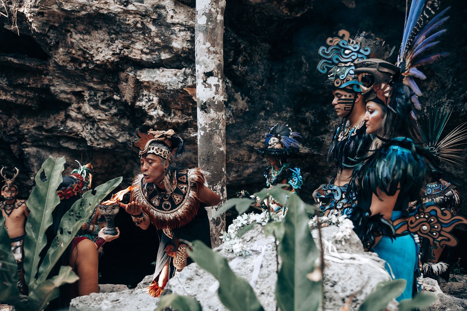 Two performers in elaborate Mayan ceremonial costumes with feathered headdresses sit among ancient stone ruins.