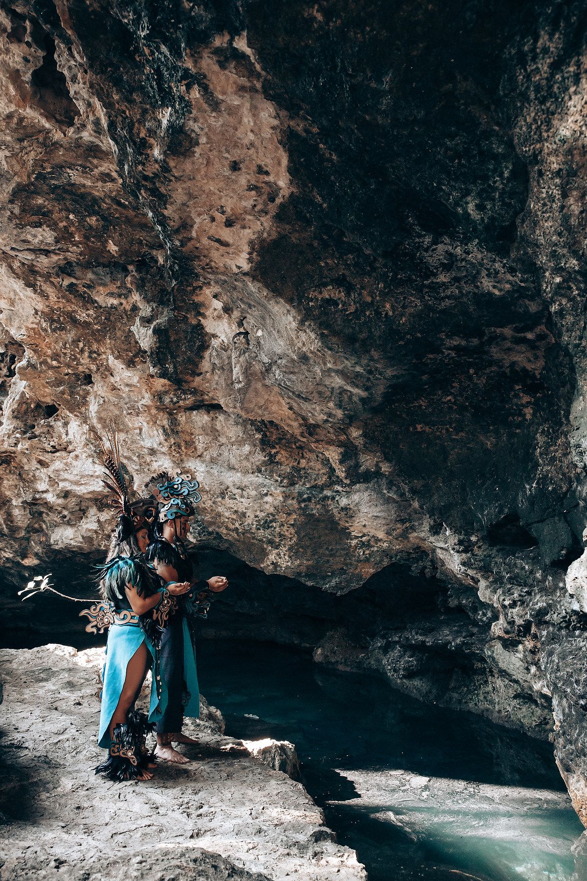 Two people in Mayan ceremonial feathered costumes stand together on rocks overlooking turquoise water in a cave.