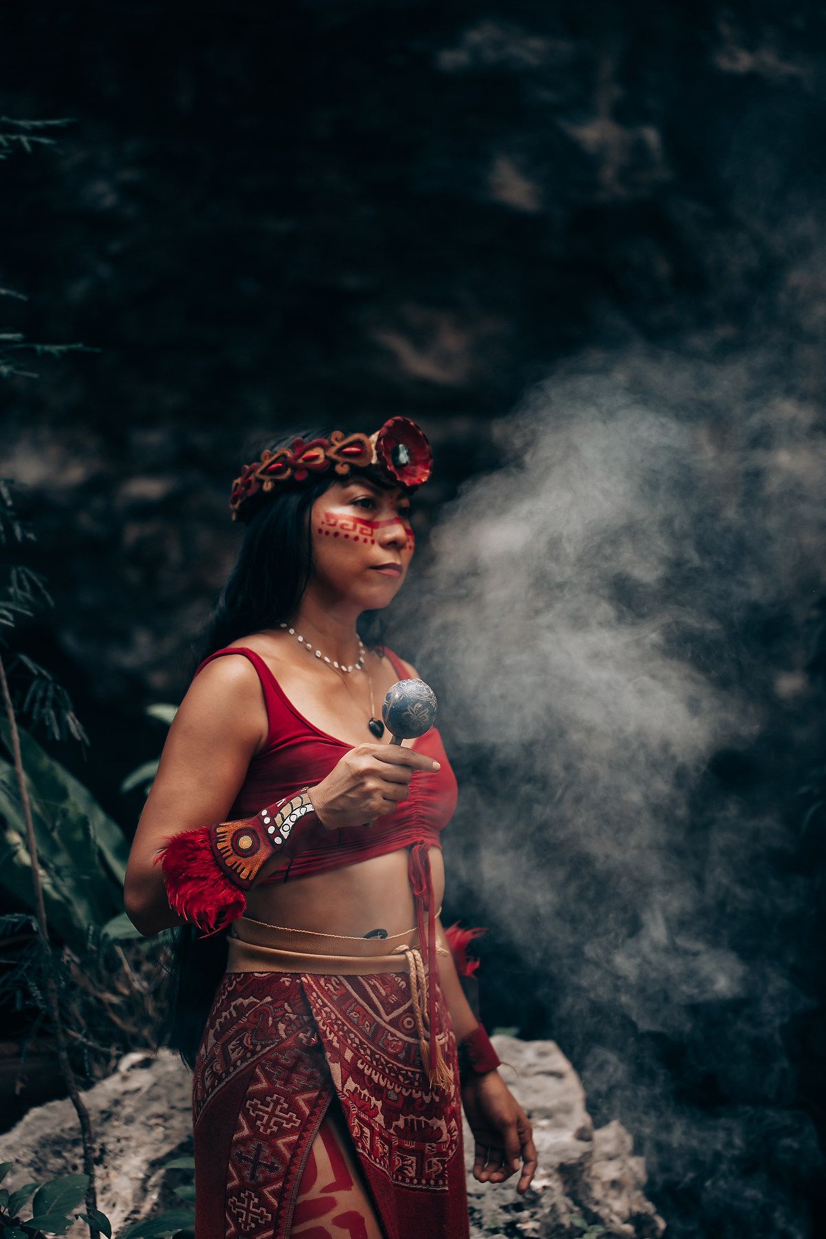 Woman in red Mayan ceremonial attire with face paint holding rattle amid mystical smoke outdoors.