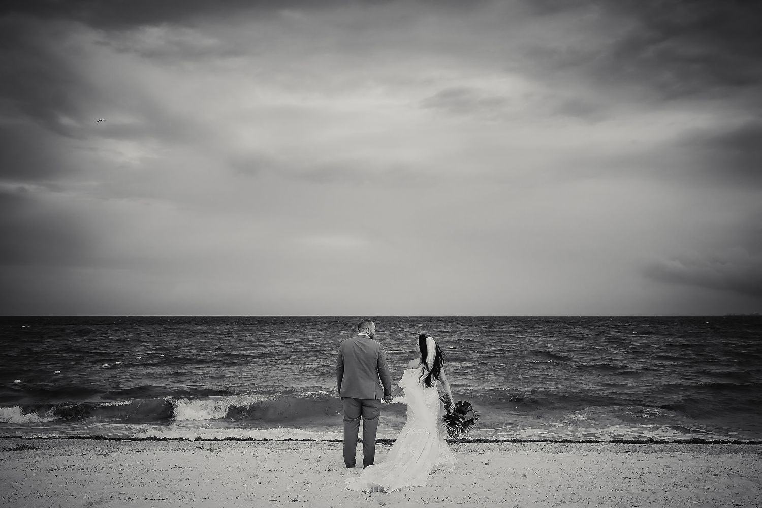 Black and white photo of a bride and groom holding hands on a beach shoreline under dramatic cloudy skies.