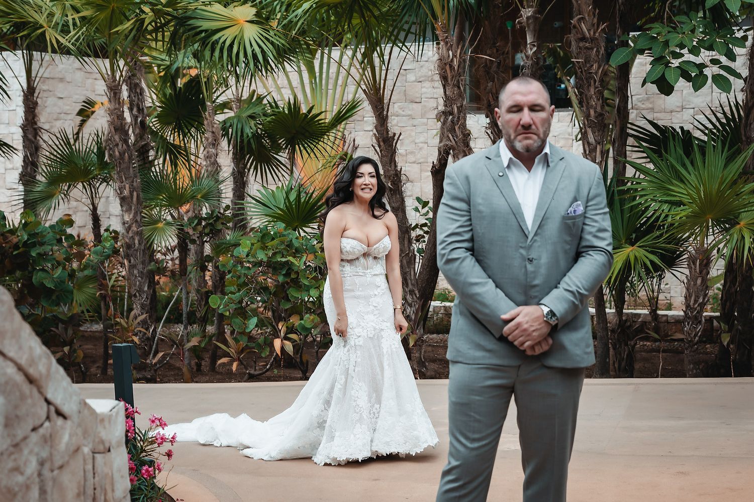 Bride in mermaid wedding gown stands behind groom in gray suit for first look reveal outdoors.