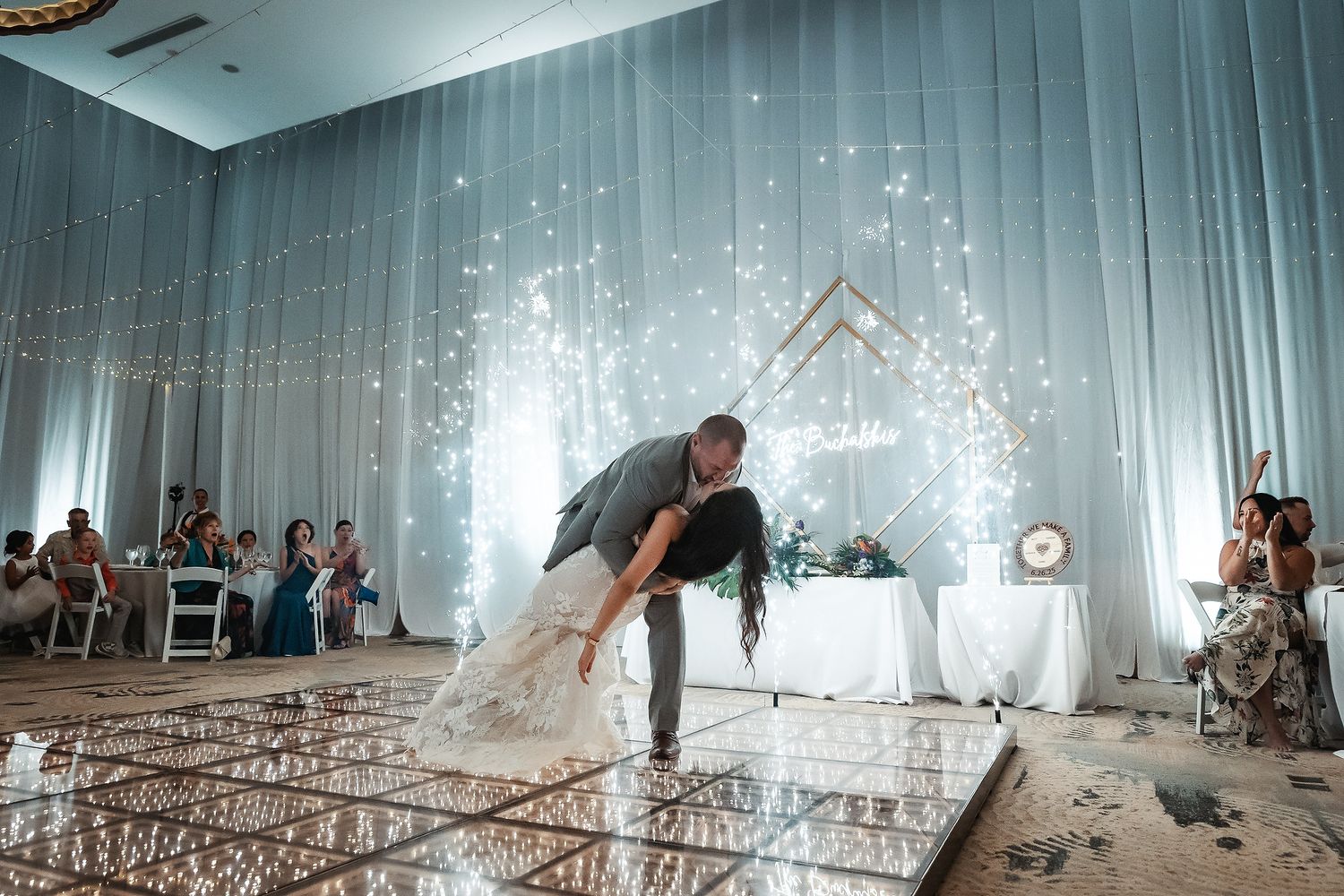 Bride and groom share romantic dip on mirrored dance floor at wedding reception with fairy light decor.