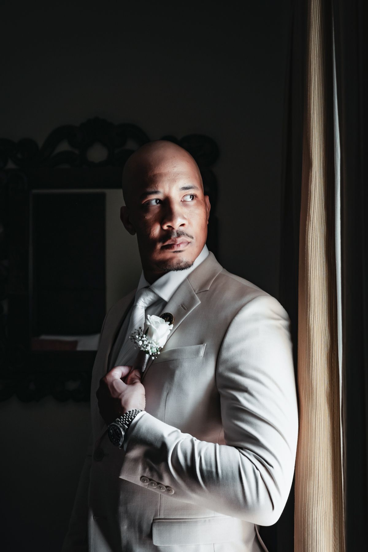 Groom in beige suit with white boutonniere posing by window in dramatic lighting.