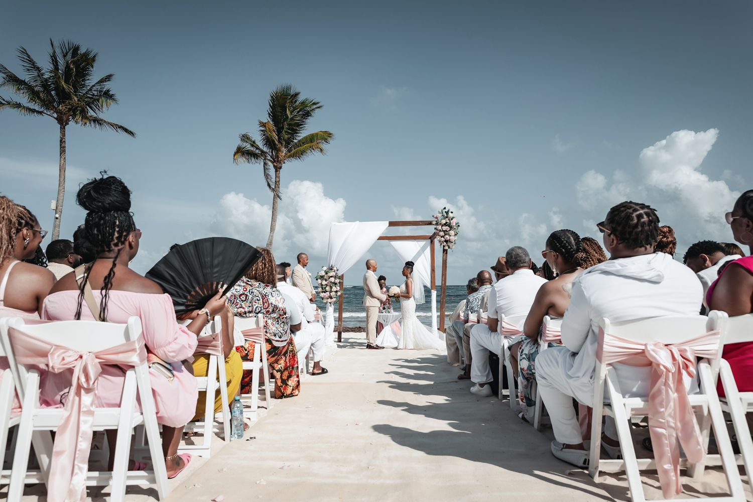 Outdoor beach wedding ceremony with guests seated in white chairs facing a floral arch under a blue sky with palm trees.