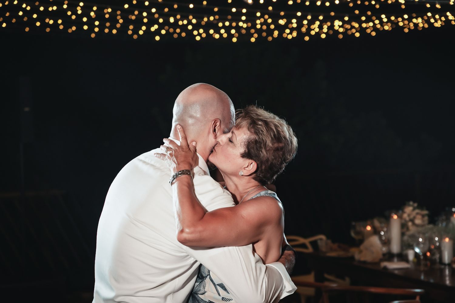 Older couple sharing a romantic kiss at an outdoor evening event with warm string lights glowing in the background.