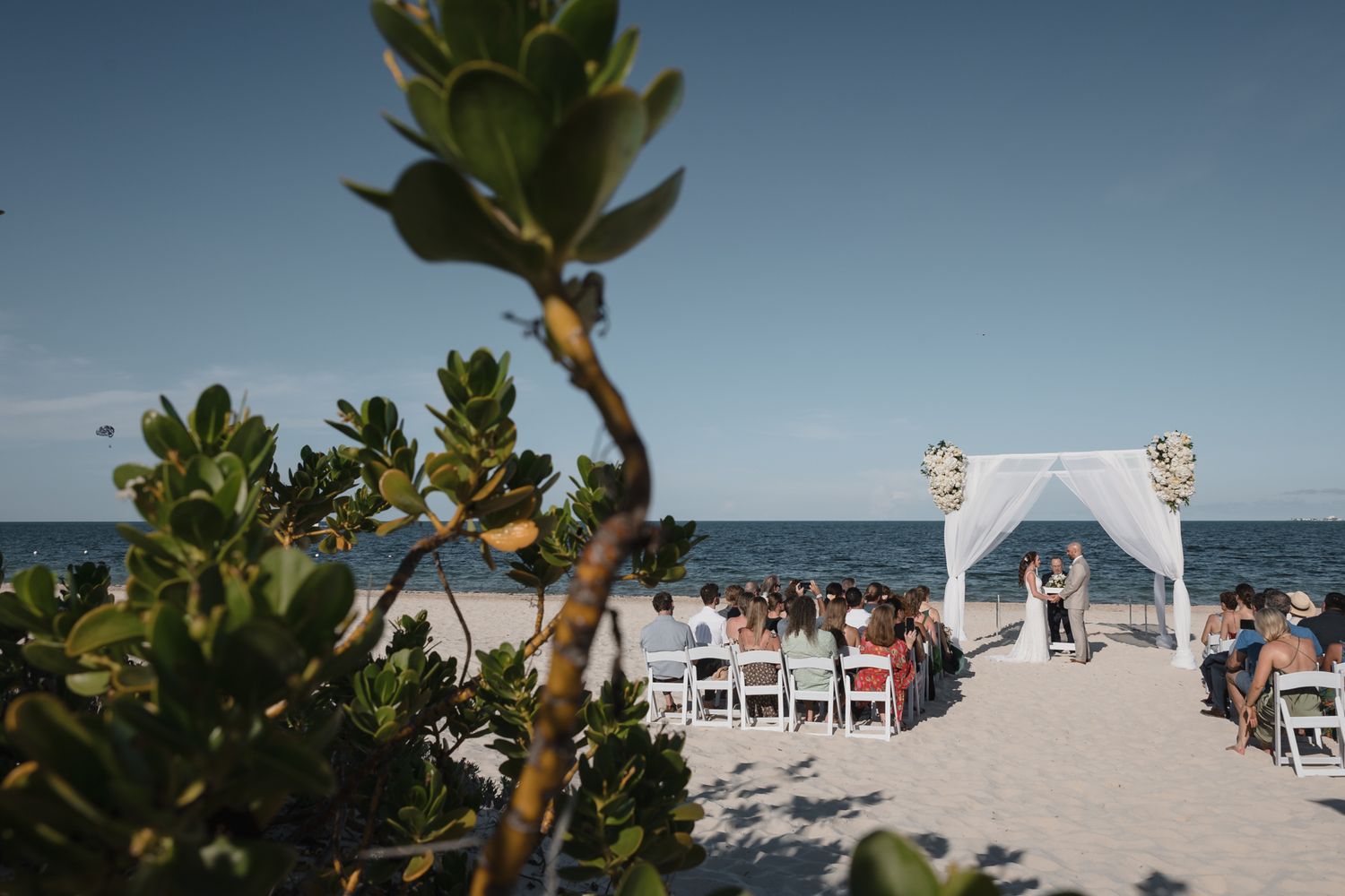 Beach wedding ceremony with white arch, floral decor, and guests seated on white chairs by the ocean under a clear blue sky.