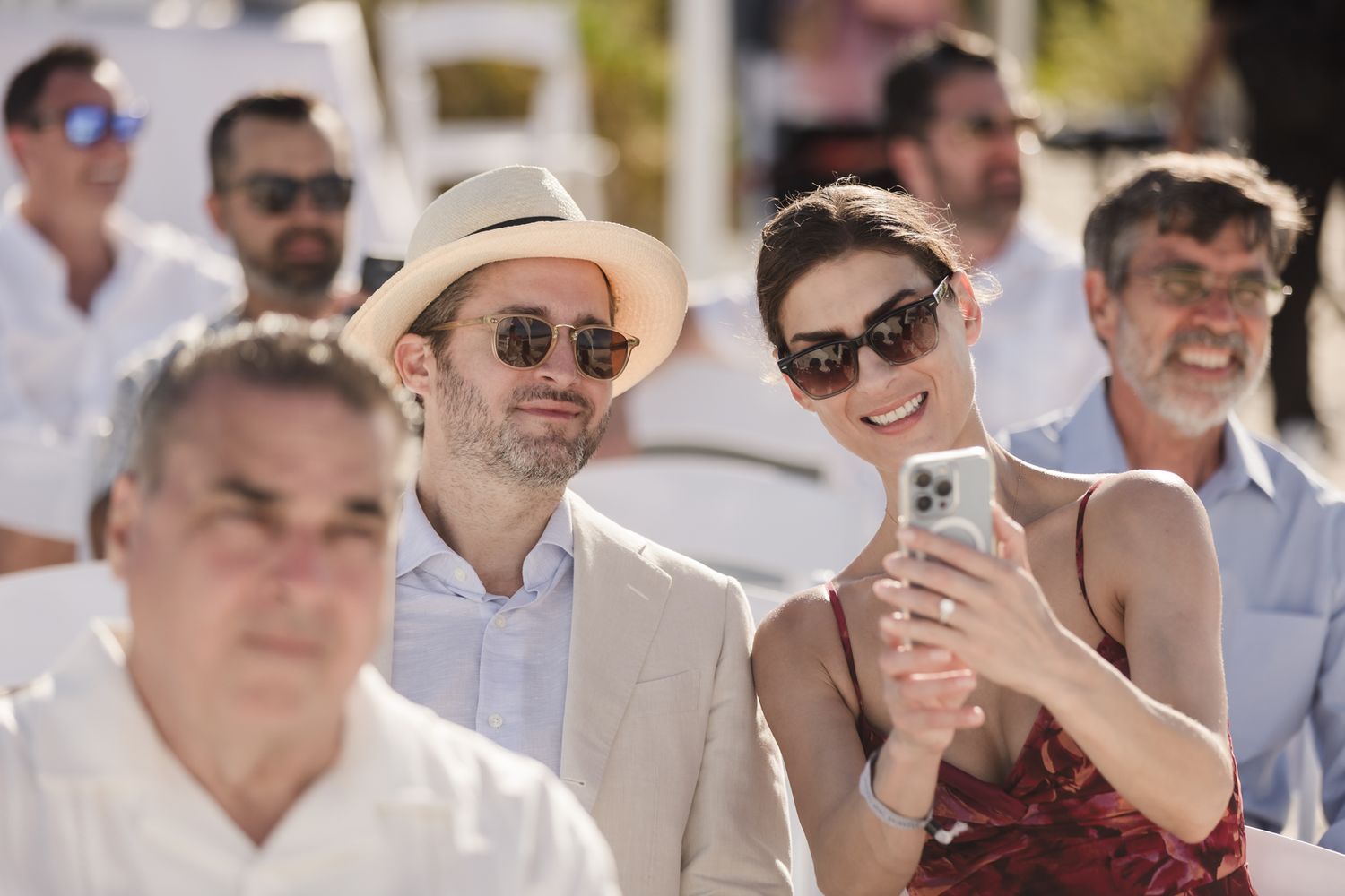 A smiling couple in summer attire takes a selfie at an outdoor wedding ceremony, surrounded by seated guests.