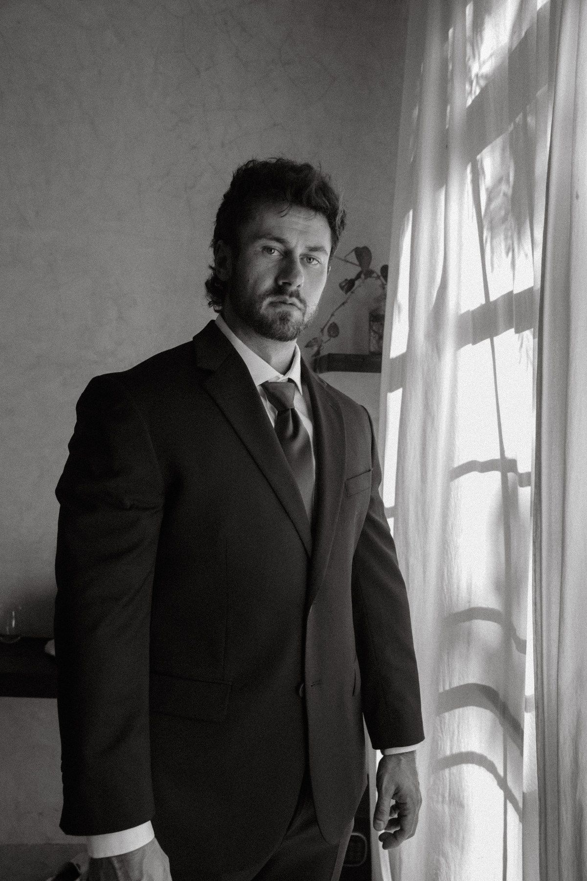 Black and white portrait of a bearded man in a dark suit and tie standing near a window with sheer curtains.