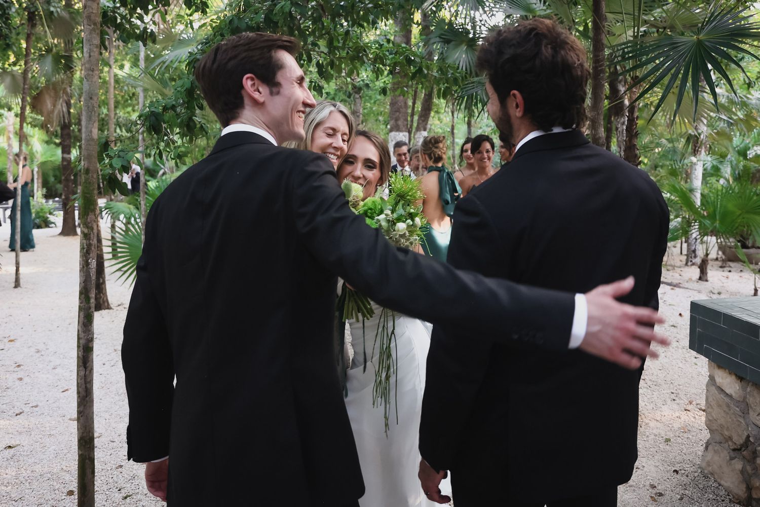 Wedding guests in black suits congratulate a smiling bride in a white gown outdoors among lush green trees.
