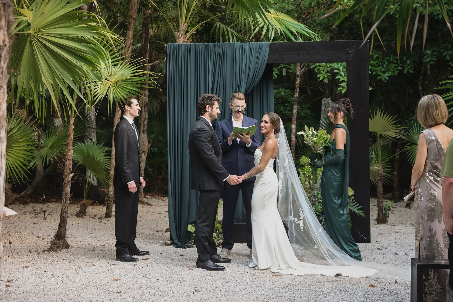 Bride and groom exchange vows under elegant dark floral arch surrounded by tropical palms.