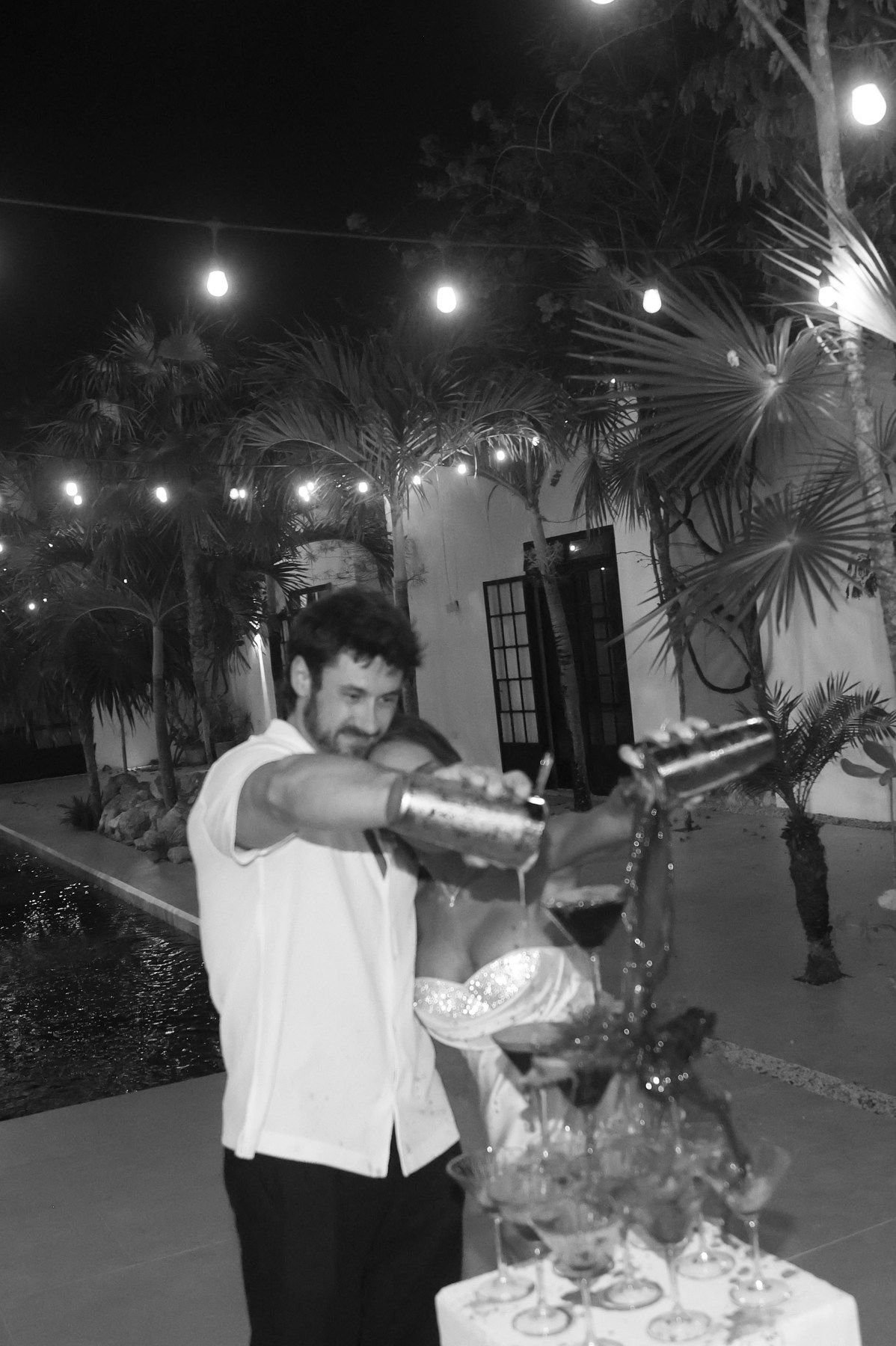 Bartender pouring cocktails at an outdoor wedding reception by a pool at night.