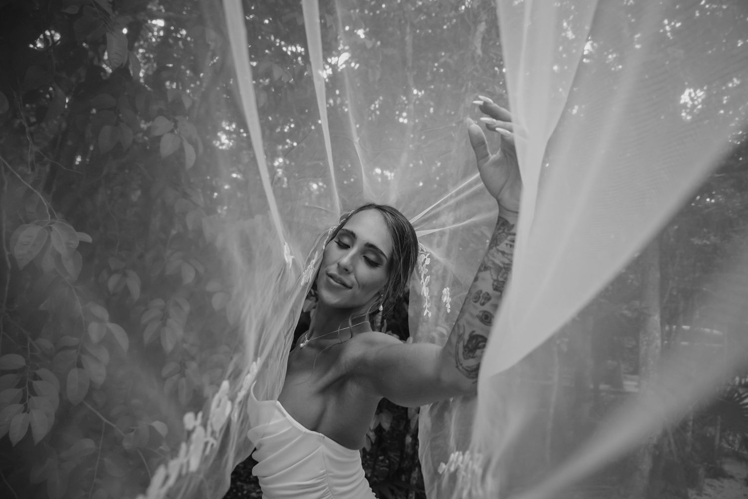 Black and white photo of a bride in a white dress holding a sheer veil outdoors.