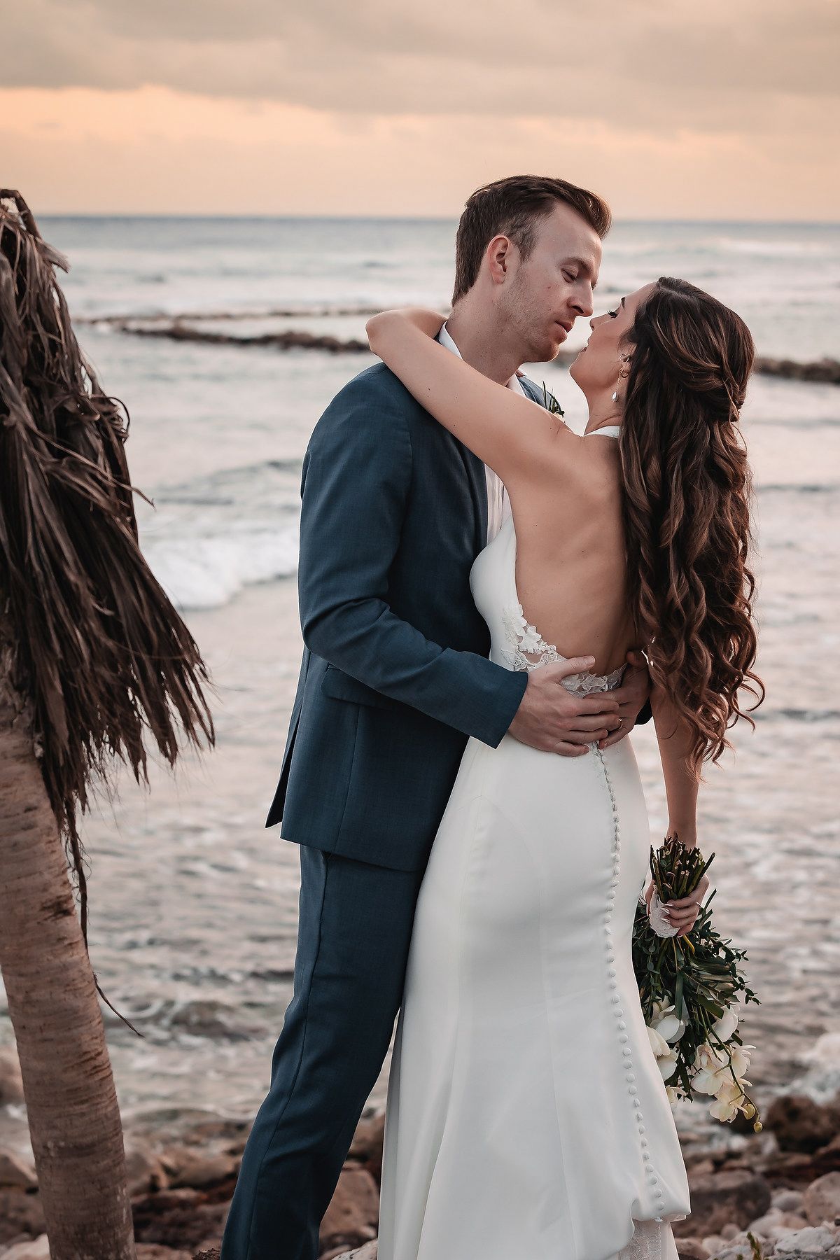 Bride and groom share romantic kiss by ocean at sunset, bride holds bouquet in backless gown.