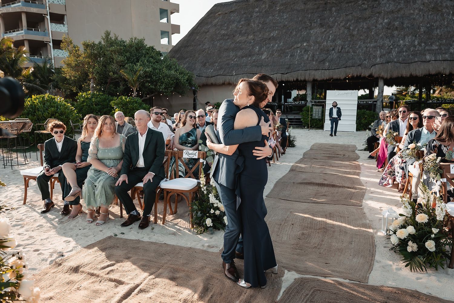 Two people embrace at an outdoor beach wedding ceremony with guests seated on both sides under a thatched roof.