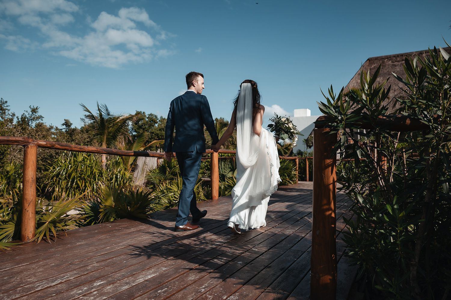 Bride and groom walking hand in hand on a wooden deck surrounded by tropical greenery under a blue sky.