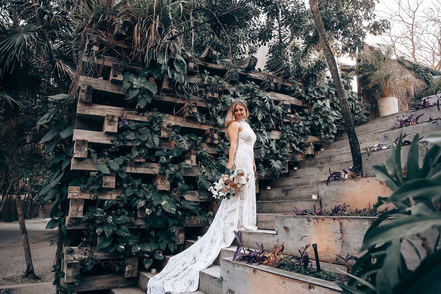 Bride in white gown with train poses on steps beside rustic wooden wall covered in lush green ivy outdoors.