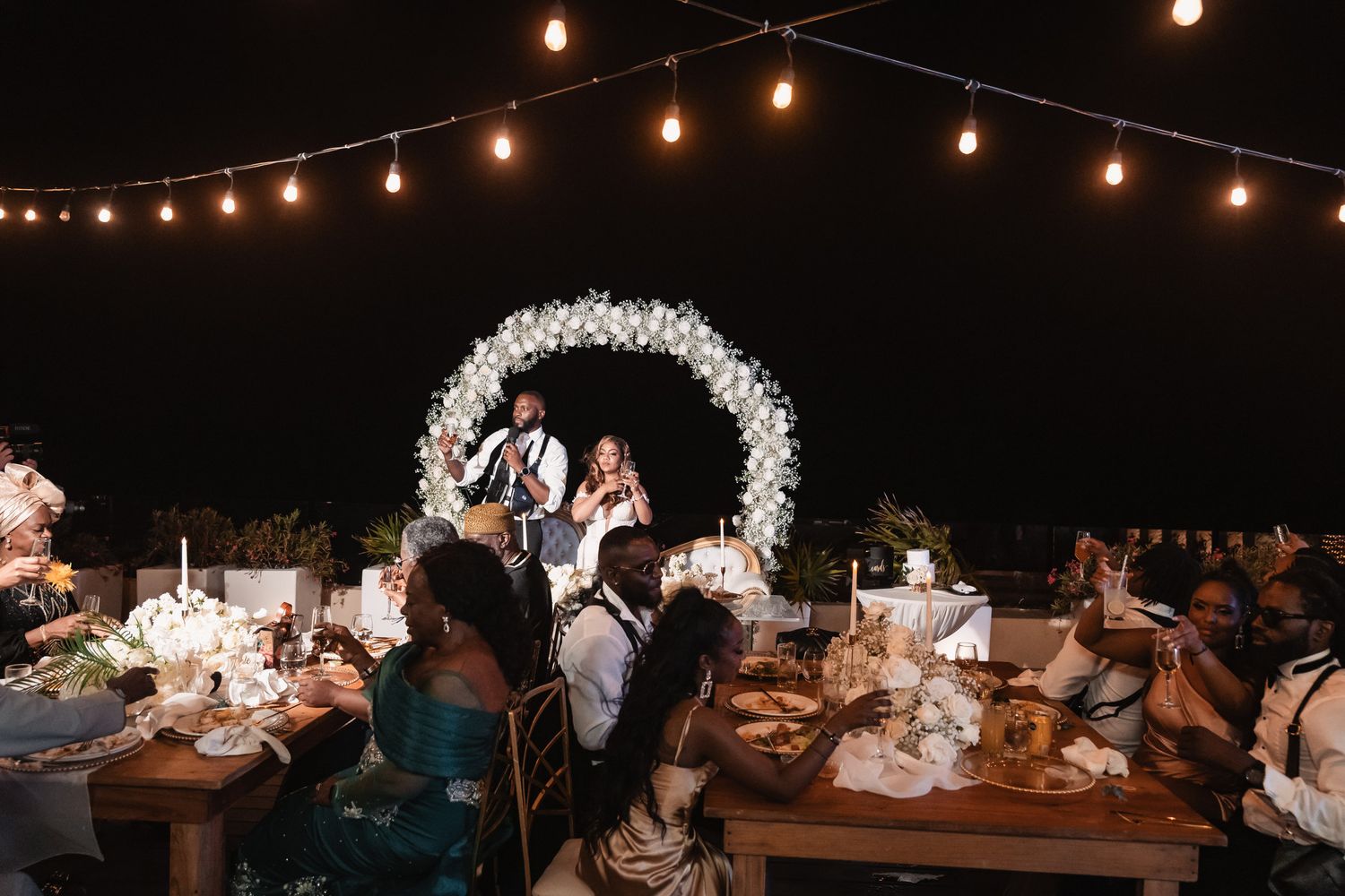 Newlyweds seated at sweetheart table under floral arch and string lights at elegant outdoor night reception.