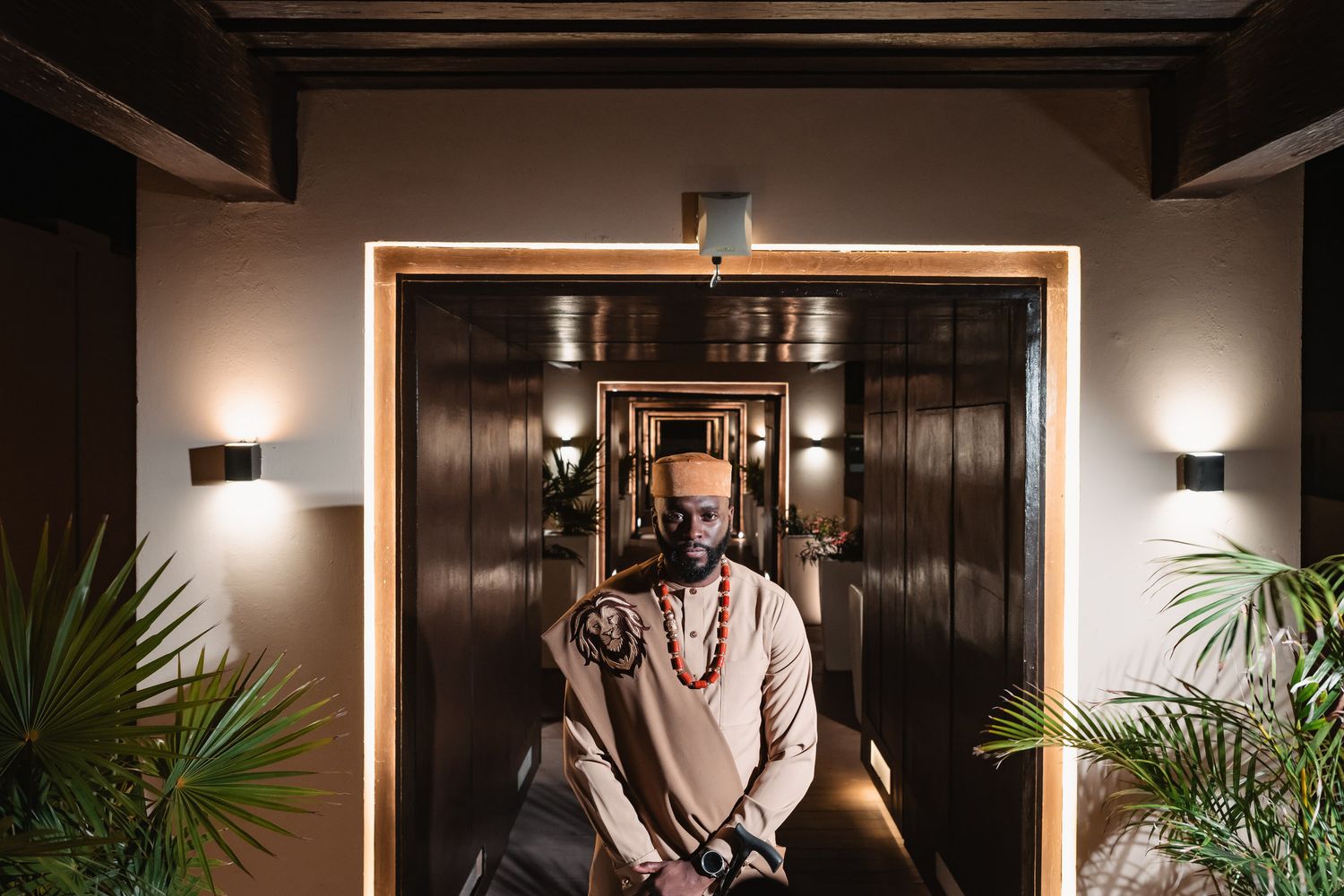 Man in traditional African ceremonial attire seated regally in a dramatic doorway with warm lighting.