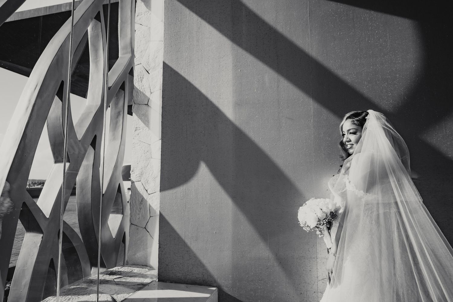 Black and white portrait of bride holding bouquet beside curved wooden architectural detail with shadows.
