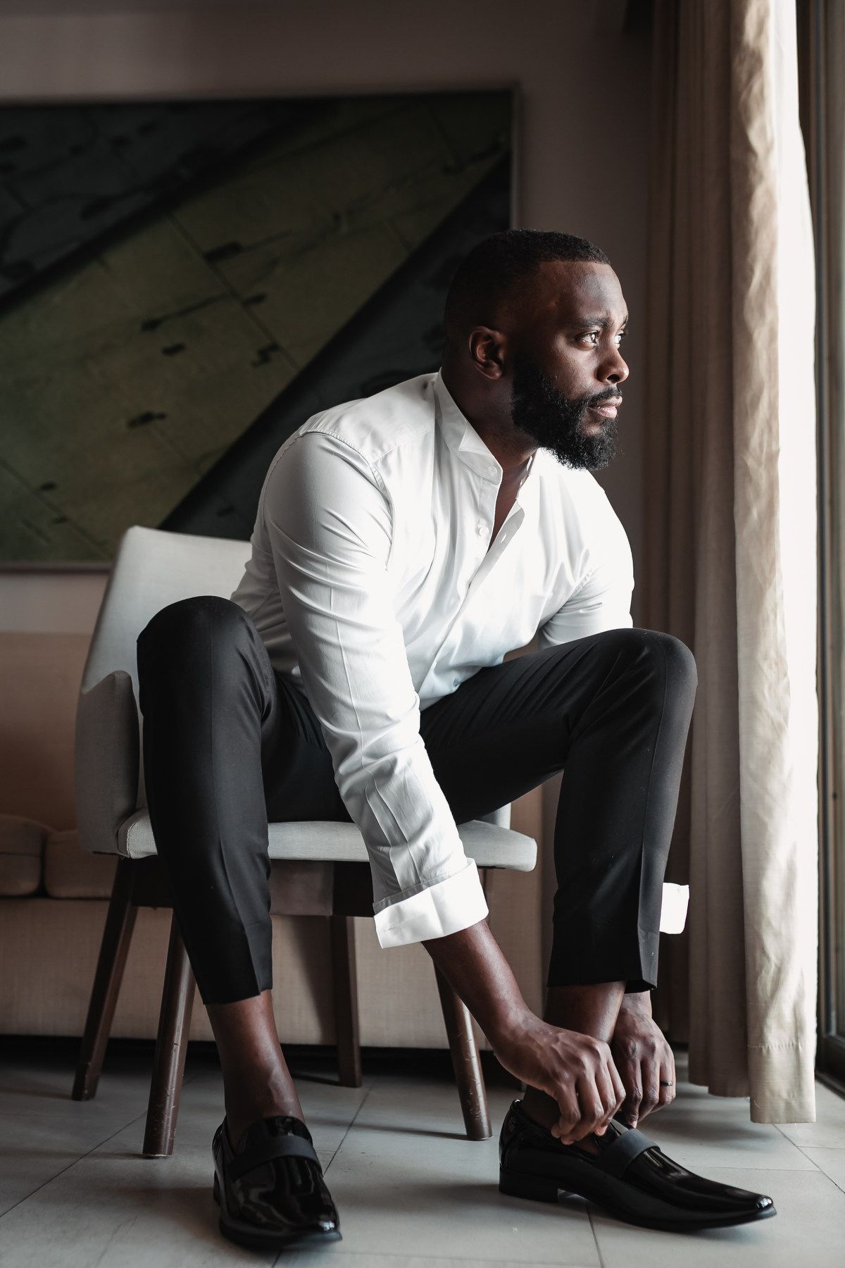 Well-dressed Black man in white shirt and black pants tying dress shoes while seated in a modern chair.