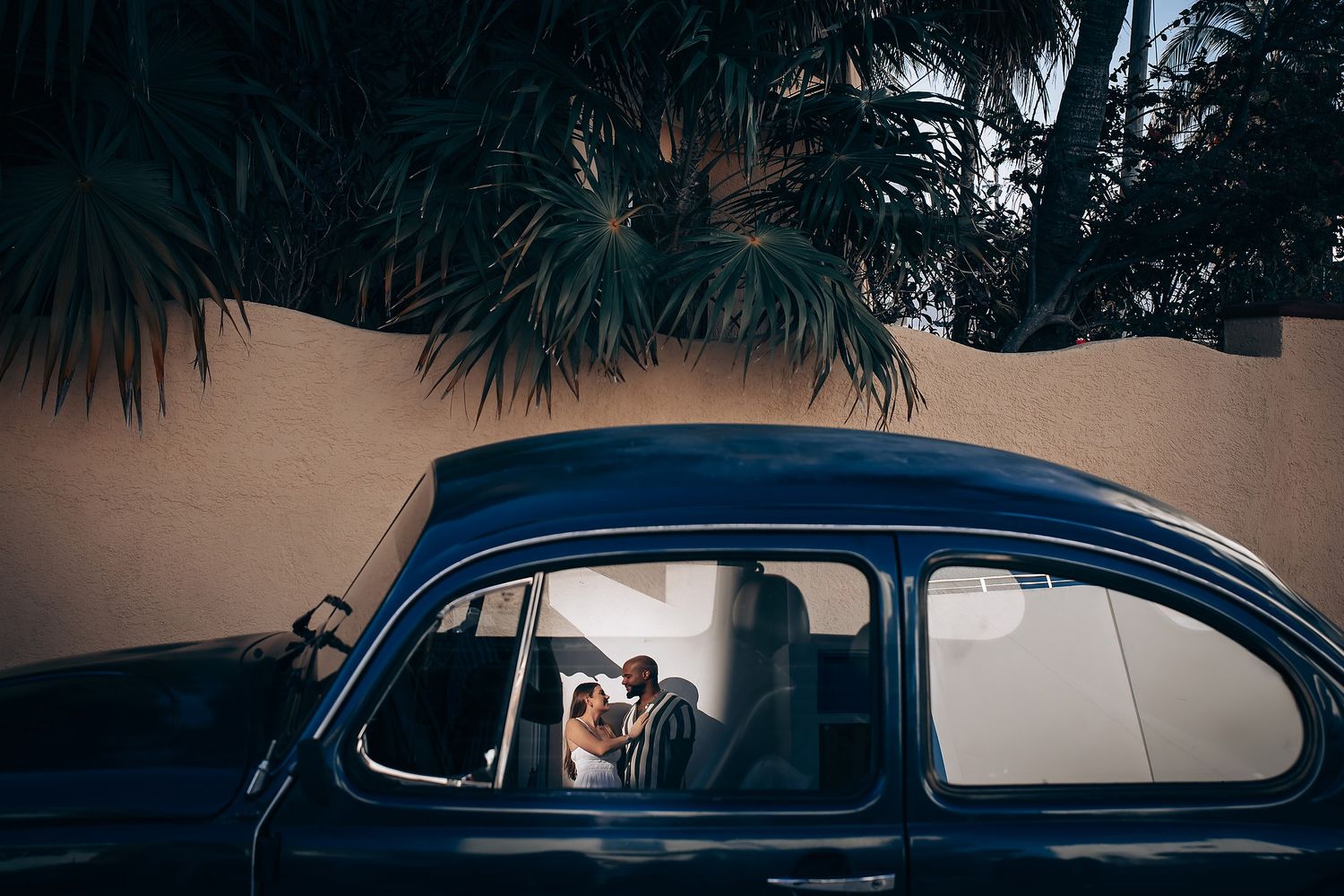Romantic couple sharing a tender moment inside a vintage blue classic car surrounded by palm trees.
