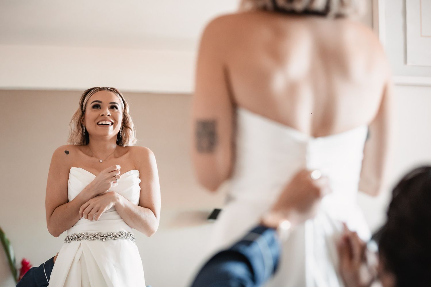Smiling bride in strapless wedding gown watching another bridesmaid get dressed in a bright room.