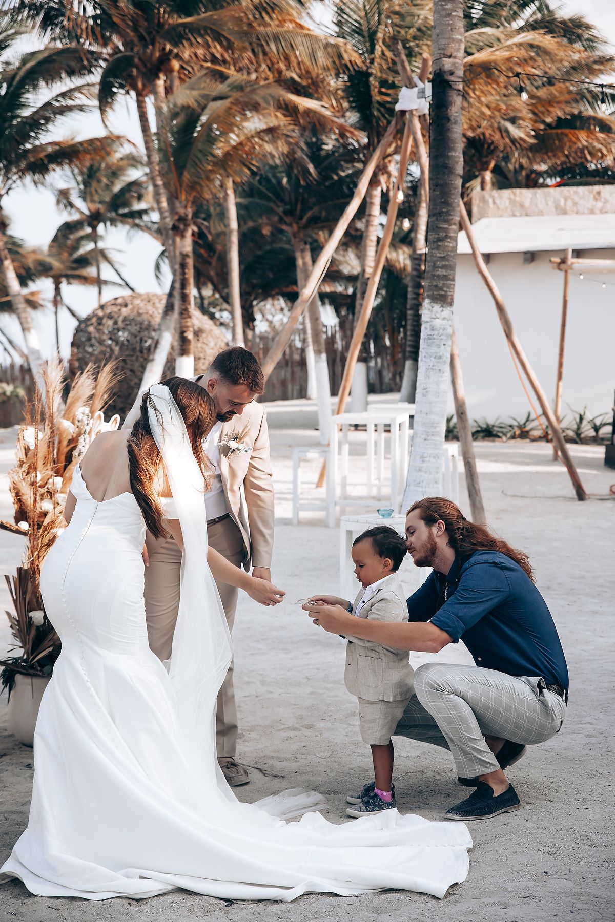 A couple sits together on a tropical beach with palm trees as the bride's white wedding dress flows in the sand.