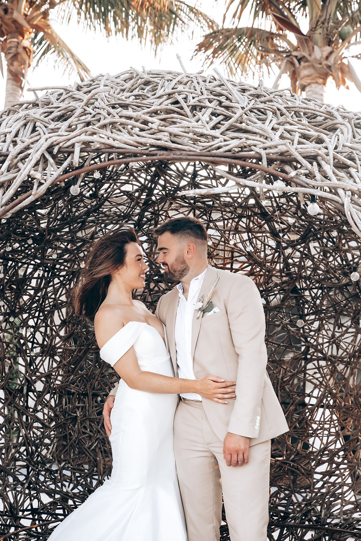 A romantic couple in coordinated white and beige outfits embracing under a natural branch archway.