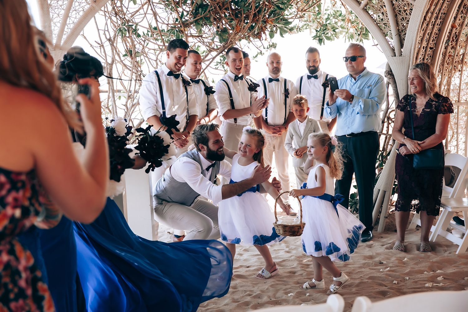 Wedding party and flower girls gather under an ornate archway for photos.