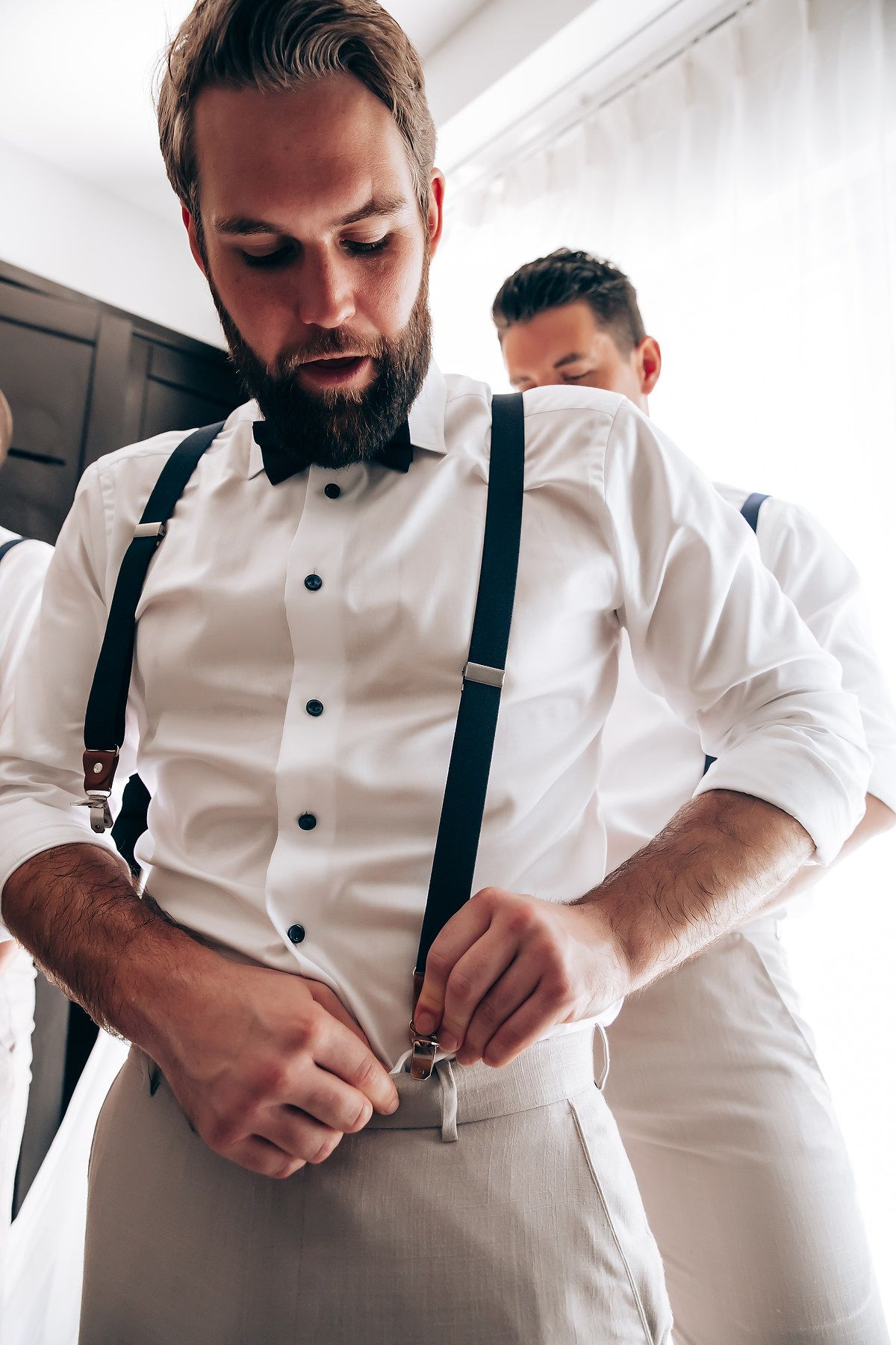 Two men getting dressed in formal attire with white shirts and black suspenders preparing for an event.