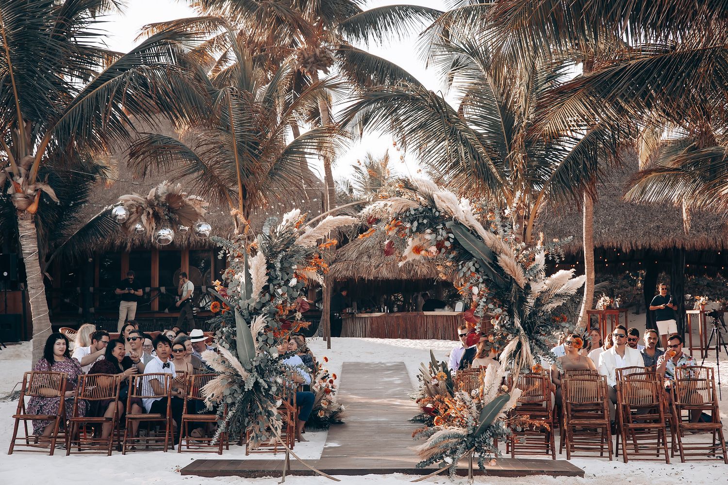 Tropical beach wedding setup with palm trees and wooden chairs arranged on sandy shore.