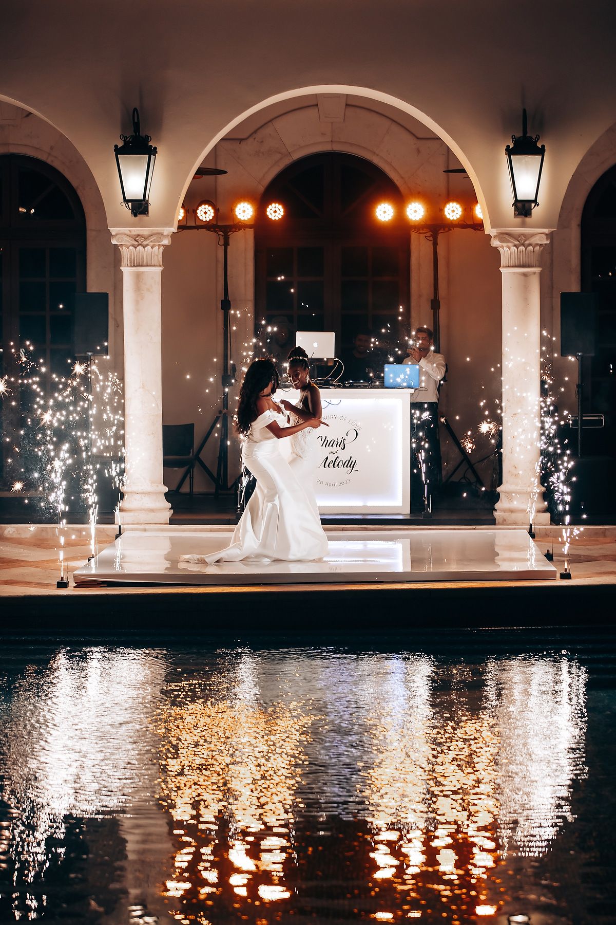 Nighttime wedding celebration by pool with illuminated arches and sparkling lights reflecting on water.