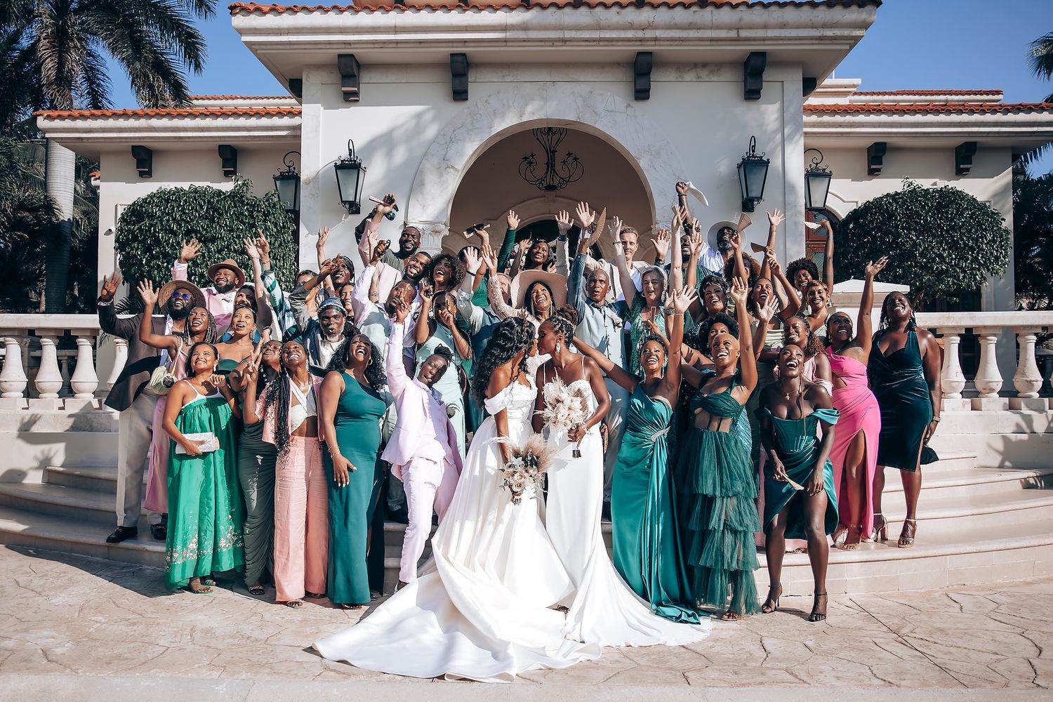 Large wedding party in emerald and neutral dresses posing at Mediterranean venue.