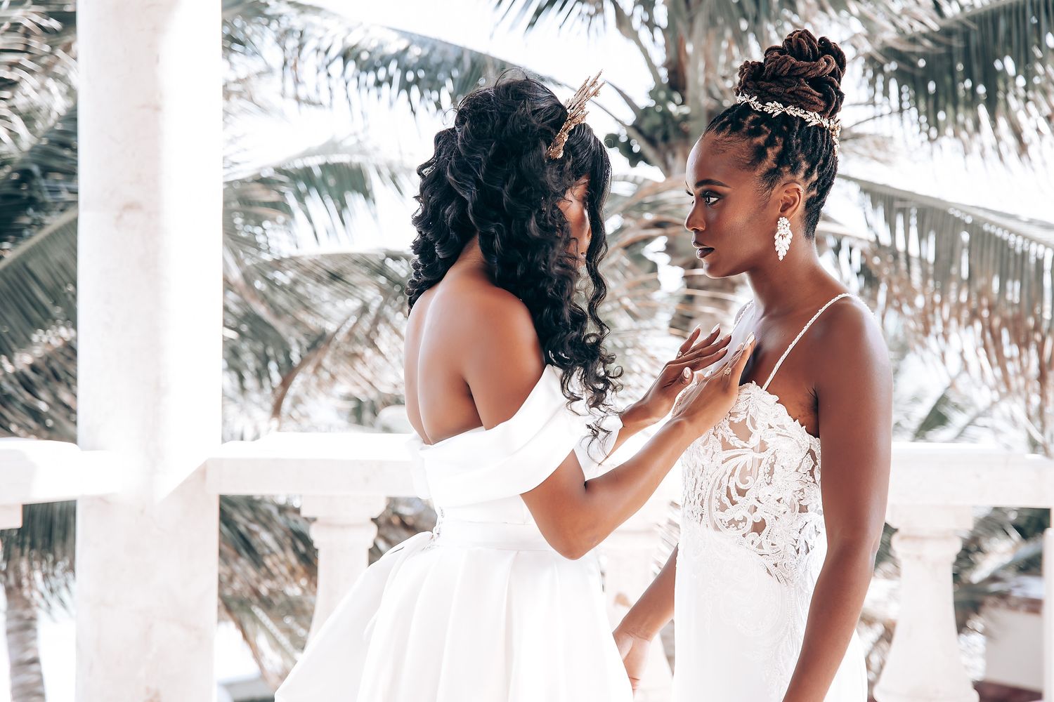 Two people in white wedding dresses share an intimate moment on a tropical balcony.