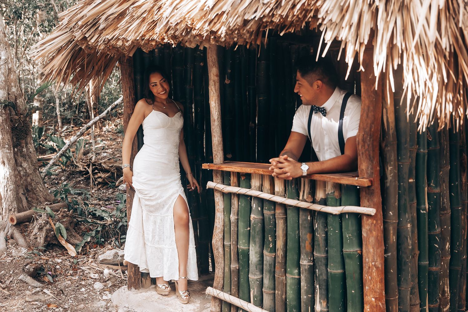 A couple in casual attire poses together in front of a rustic thatched-roof hut in a tropical setting.