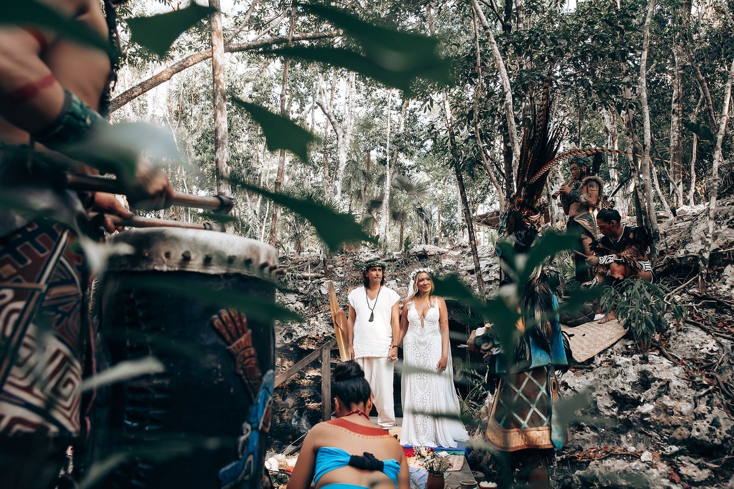 People gather outdoors for a celebration in a lush forest setting with natural sunlight filtering through trees.